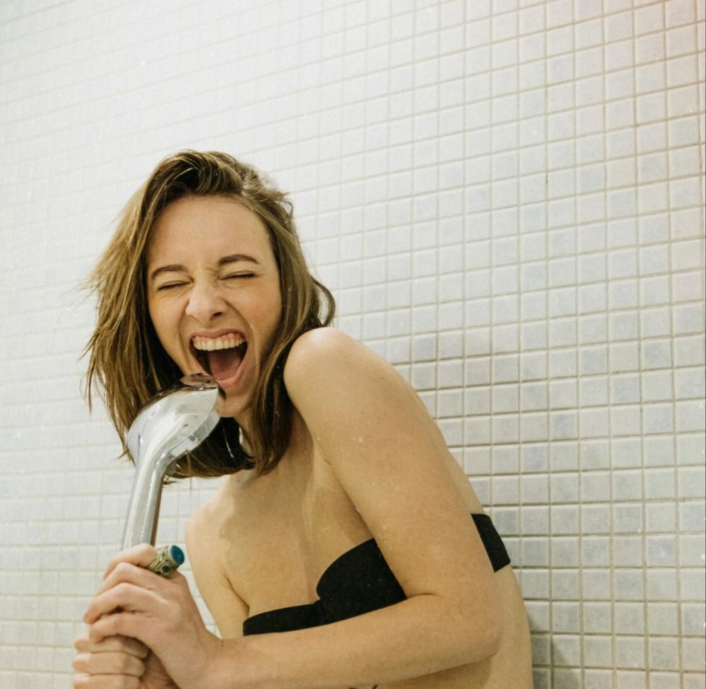 A joyful woman sings with a shower head in a bathroom, showcasing her playful spirit.